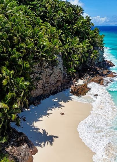 A secluded white-sand beach cove framed by granite boulders and palm forest, with turquoise waves rolling in